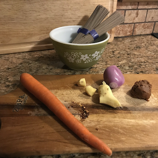 a cutting board with carrot, shallot, ginger, chile pepper, miso paste, and a bowl of dry soba noodles in the background